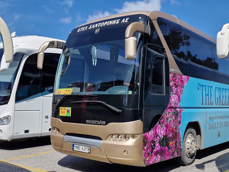 A striking KTEL Santorinis Neoplan coach wrapped in a vibrant bougainvillea flower livery, advertising 'The Greek' daily service to Fira.