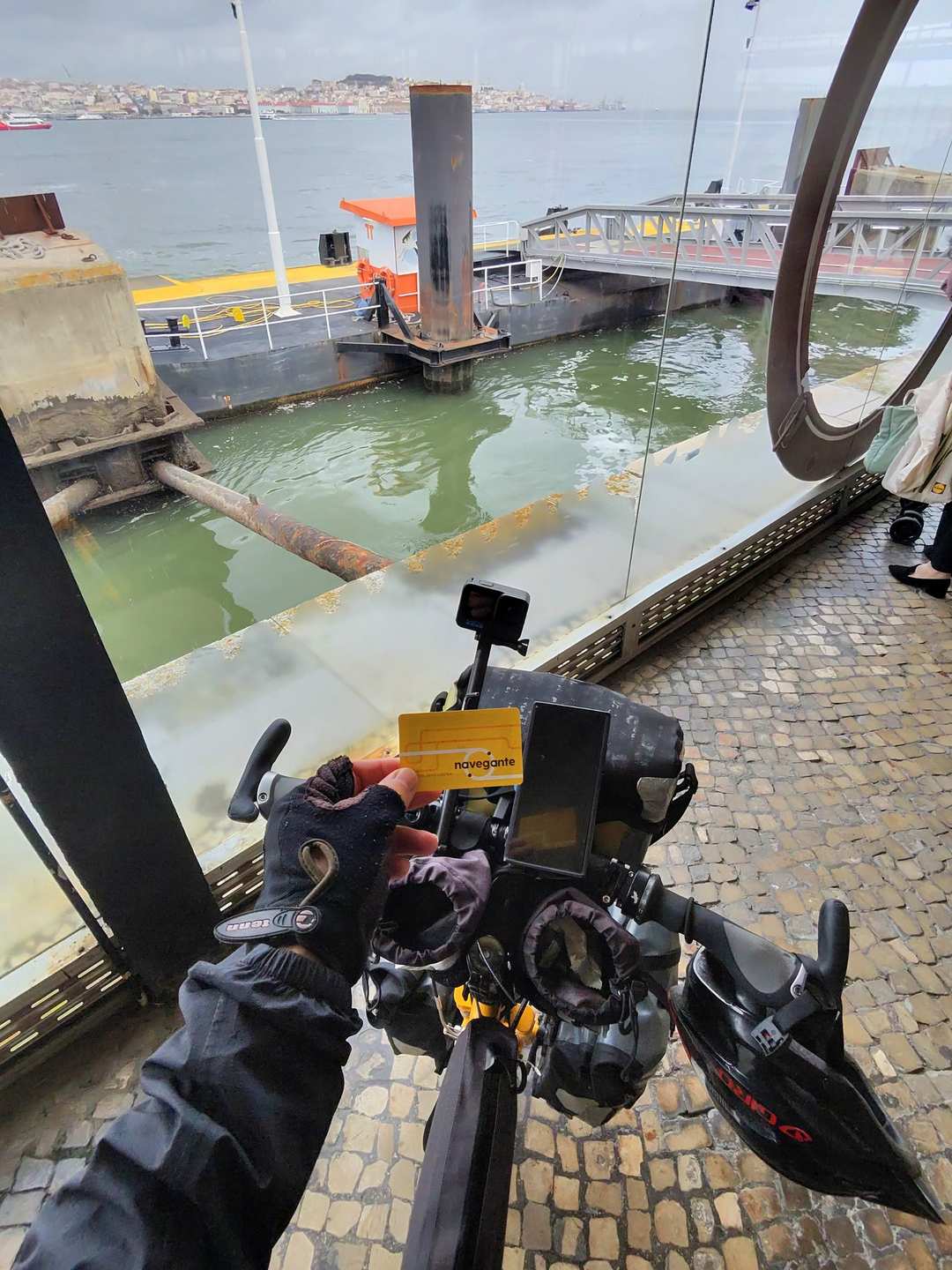 View from bicycle handlebars approaching a yellow ferry terminal building on a grey rainy day with Lisbon across the water