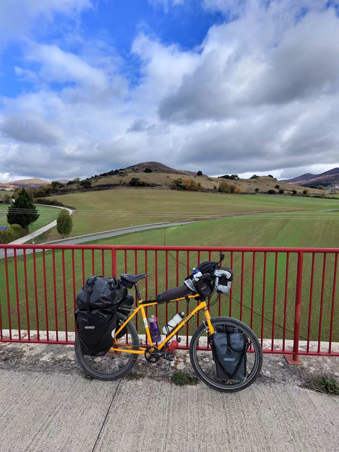 A loaded yellow touring bicycle leaning against red railings with green hills and mountains under cloudy skies